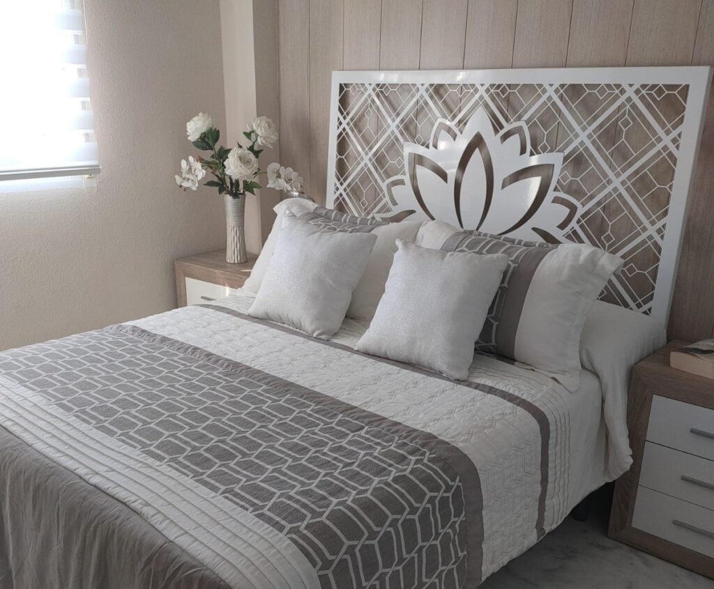 Cabecero de cama de diseño en metal lacado en blanco, fabricado a medida mediante corte láser con un elegante motivo central de flor de loto sobre un patrón geométrico de rombos. Instalación decorativa en el dormitorio principal de una vivienda en la zona de Churriana, Málaga.