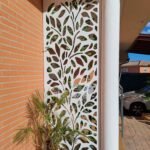 Panel de privacidad y ocultación metálico lacado en blanco, con un diseño orgánico de hojas fabricado mediante corte láser. Instalación a medida funcionando como celosía exterior decorativa para dar sombra y separar el espacio junto a un pilar y una fachada de ladrillo visto en una vivienda en Madrid.