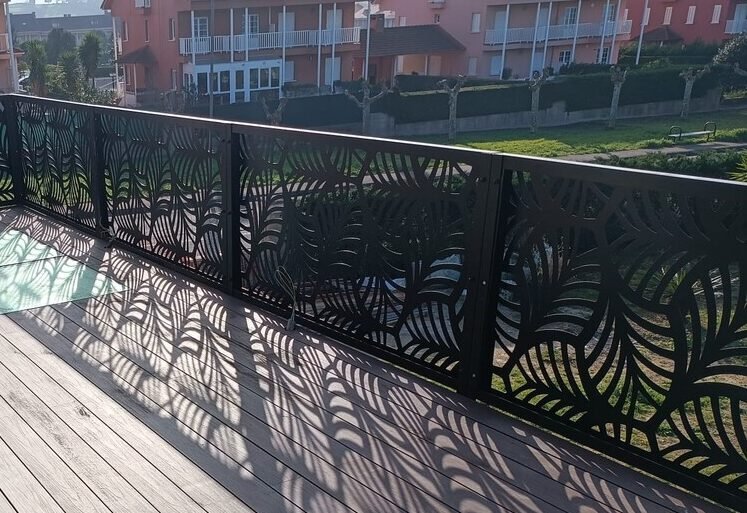 Barandilla metálica de exterior en color negro para terraza con suelo de madera, con diseño botánico de hojas en corte láser proyectando sombras, instalada en Galicia.
