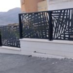 Valla perimetral metálica en color negro con diseño botánico en corte láser, instalada sobre un muro blanco exterior con montañas de fondo en Huelva.