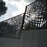 Valla metálica perimetral en color negro con diseño botánico de hojas en corte láser, instalada sobre un muro blanco de exterior a contraluz con árboles de fondo.