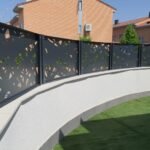 Valla perimetral metálica en color negro con diseño abstracto en corte láser, instalada sobre un muro curvo blanco en el jardín exterior de una vivienda de lujo en Madrid.