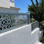 Valla perimetral metálica en color gris con diseño geométrico en corte láser y remate superior con puntas anti escalada. Cerramiento de alta seguridad con diseño anti robo instalado sobre un muro blanco exterior en una villa en Málaga.
