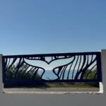 Valla perimetral de acero inoxidable en color oscuro con diseño exclusivo de cola de ballena en corte láser, instalada entre pilares blancos con vistas al mar en Valencia.