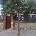 Valla perimetral metálica en color negro con diseño botánico de hojas grandes en corte láser, instalada sobre un muro de piedra y ladrillo en la fachada de una vivienda adosada en Málaga.