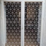 Reja metálica de doble hoja en color blanco para puertas de balcón, diseñada con un patrón geométrico continuo en corte láser. Solución de seguridad transpirable ideal para la protección de gatos y animales domésticos, instalada en una vivienda de Sevilla.
