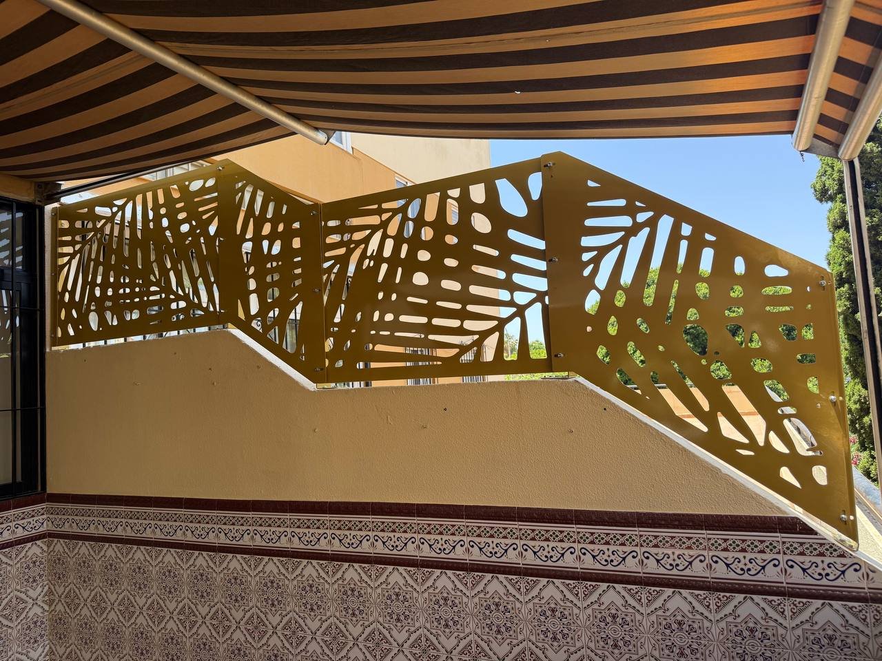 Valla metálica sobre muro escalonado en color mostaza, fabricada a medida en corte láser con diseño botánico de hojas tropicales para terraza exterior en Málaga