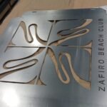 Rótulo metálico personalizado fabricado mediante corte láser con un diseño de hojas tropicales y el texto Zafiro Beach Club troquelado en la parte inferior.